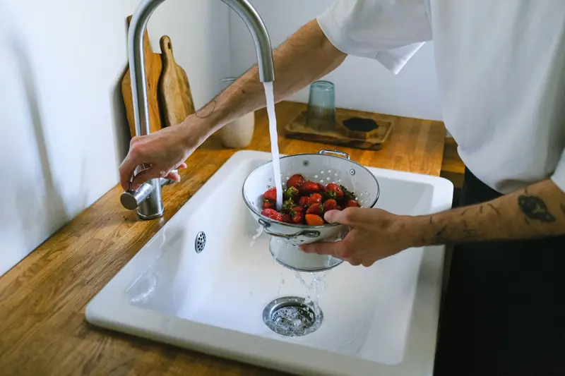 A person washes fresh strawberries in a colander under a kitchen faucet